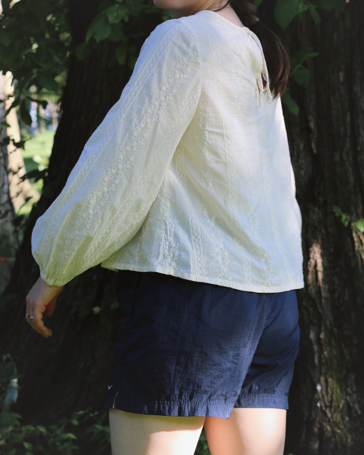 A women wearing an ivory blouse with embroidery and navy gym shorts. She's standing in front of a tree with green foliage .