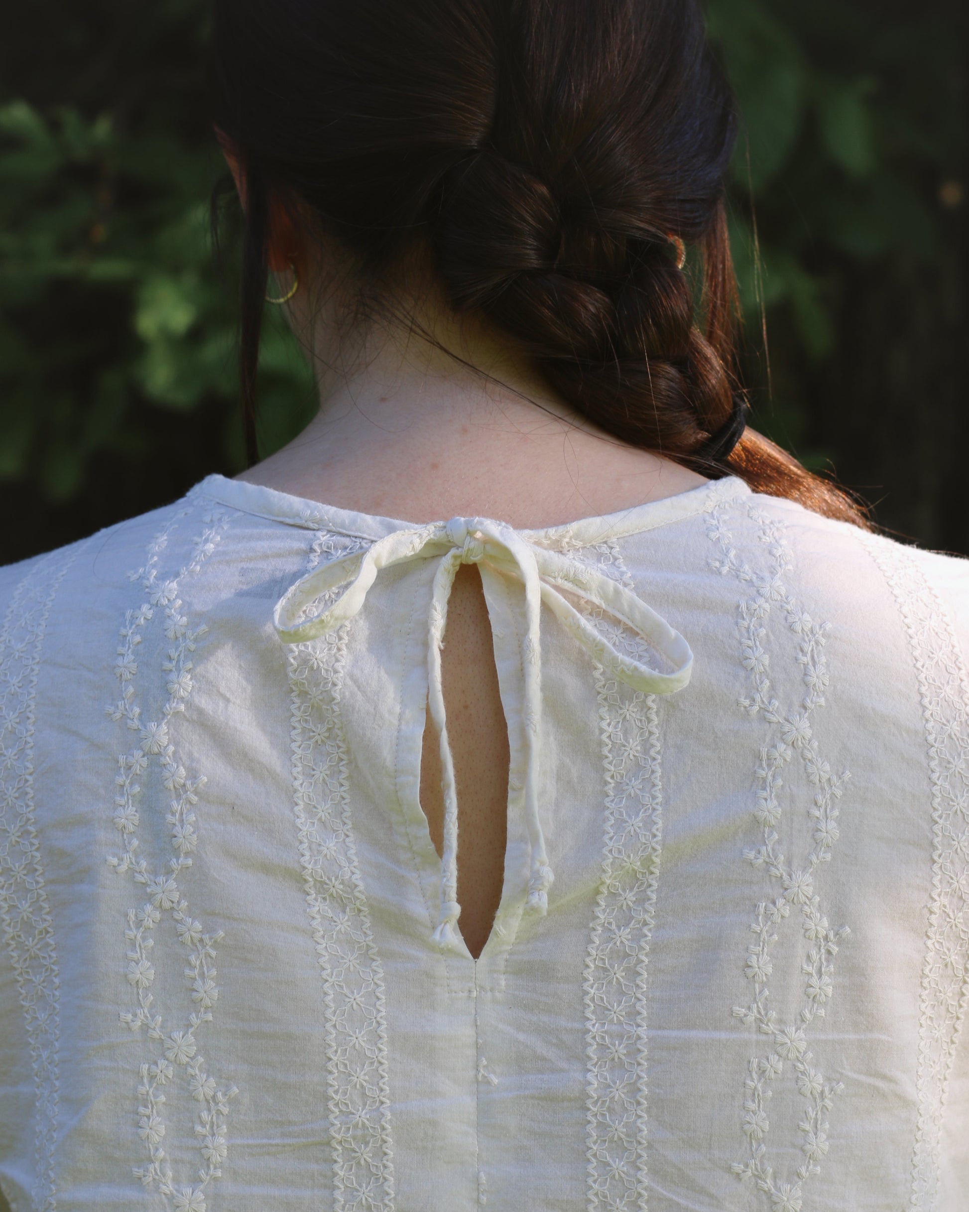 An close up image of a women wearing an ivory blouse with embroidery. The image focuses on the blouse's tie closure at center back neck.
