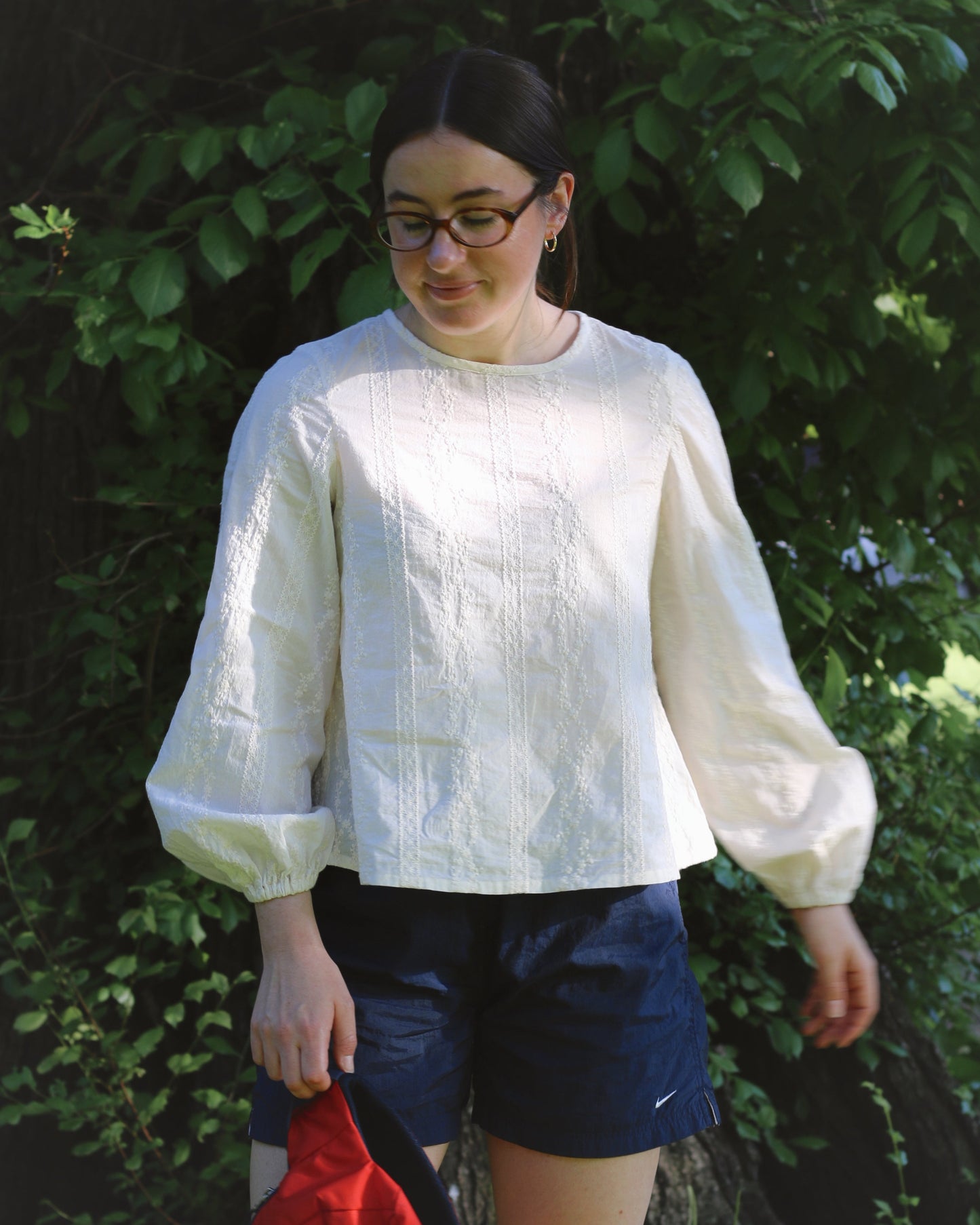 A woman wearing an ivory blouse and navy shorts. She's standing in front of a tree with green foliage.