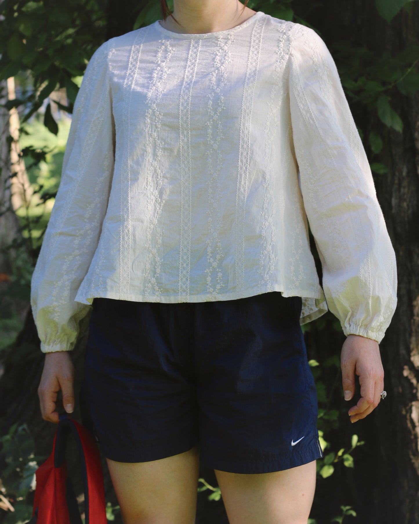 A women wearing an ivory blouse with embroidery and navy gym shorts. She's standing in front of a tree with green foliage while holding a red back in her hand.