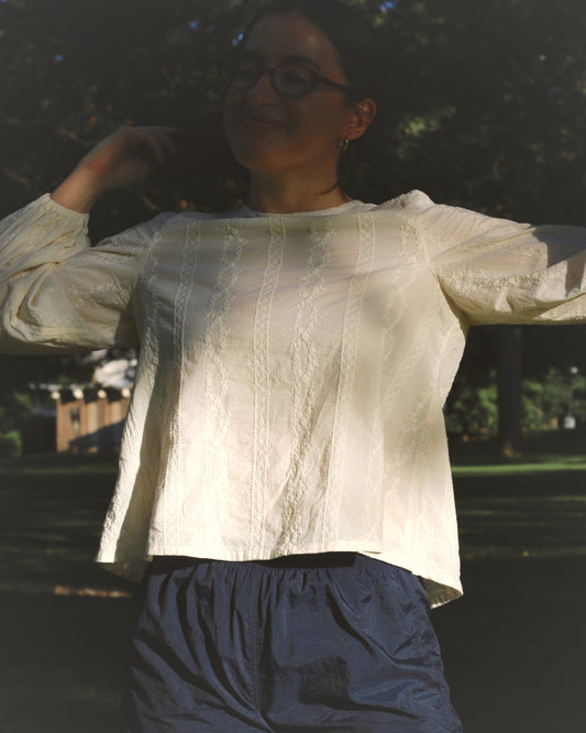 A woman wearing an embroidered ivory blouse and navy gym shorts while standing in a park.